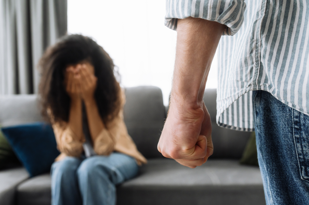 Man with a clenched fist threatening a woman, while she sits on a couch in the background covering herself with her hands, representing the concept of domestic violence.