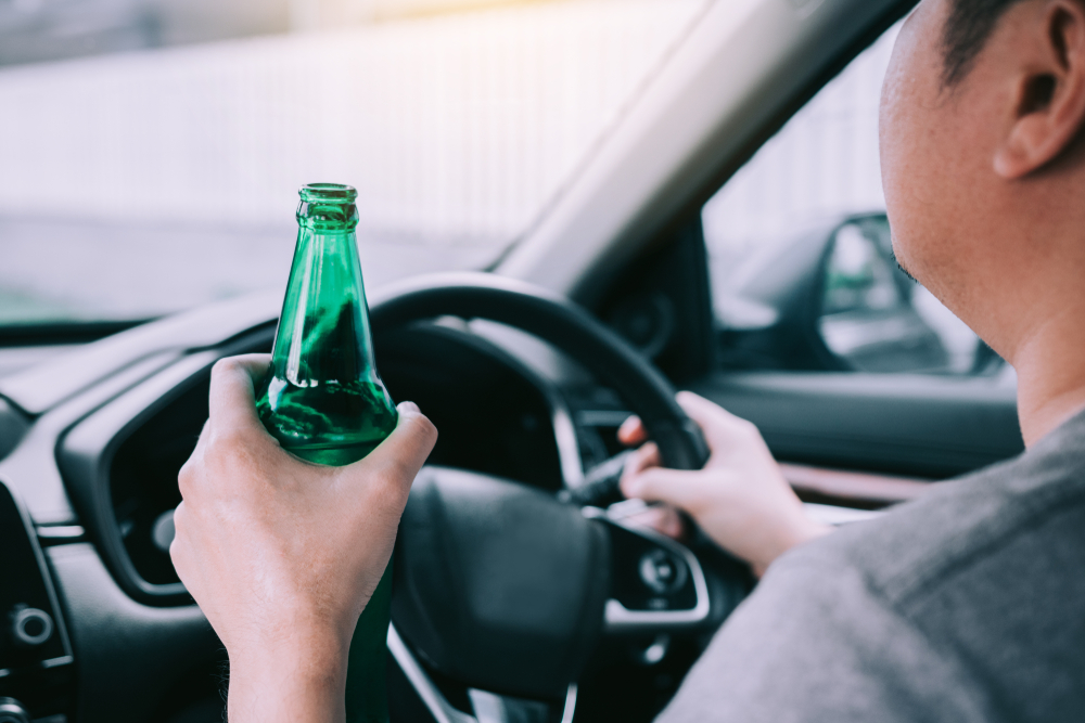 A person holds a green bottle while driving a car, emphasizing distracted or unsafe driving. The interior is sunlit, highlighting a concerning behavior.