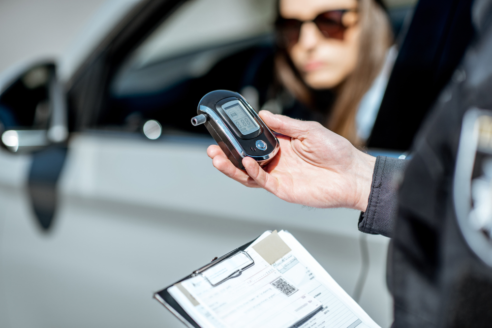A police officer holds a breathalyzer near a car with a clipboard, as a woman driver, wearing sunglasses, watches concerned from the driver's seat.