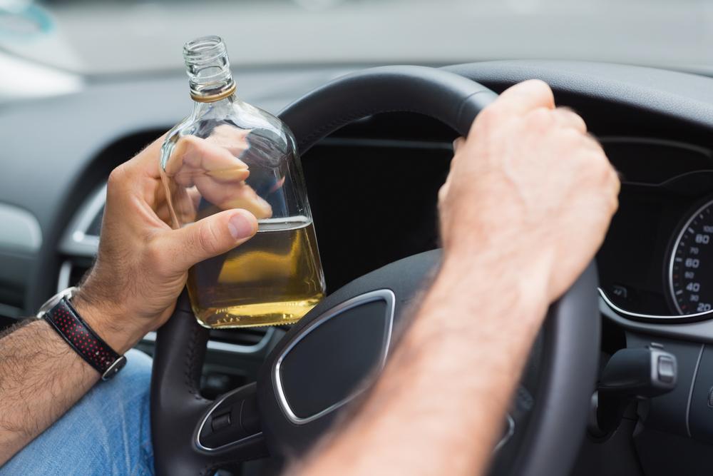A person holding a bottle of liquor while driving, gripping the steering wheel with the other hand. The image conveys a sense of danger and irresponsibility.