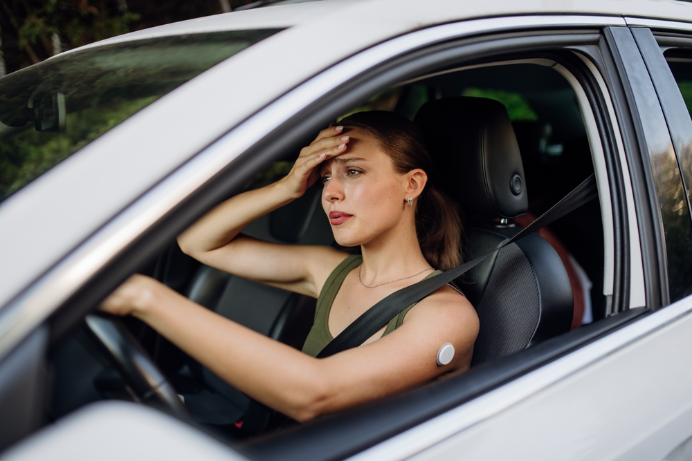 Woman driving a car with her hand on her forehead looking unwell, wearing a glucose monitor sensor on her arm