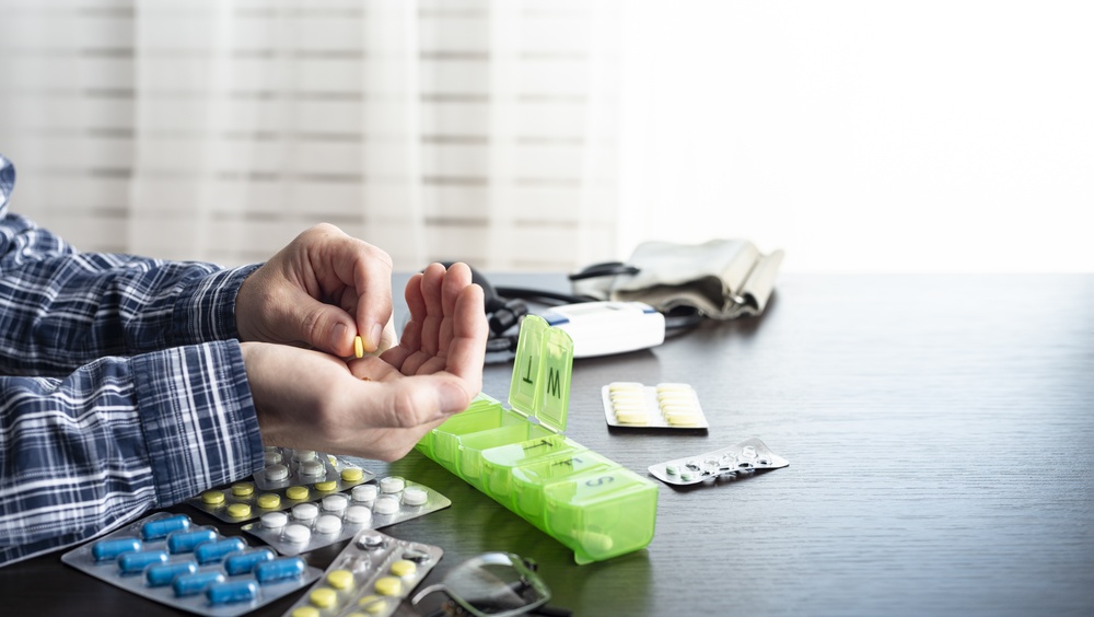 Person taking pills from a weekly pill organizer at a table with blister packs of medication and a blood pressure monitor